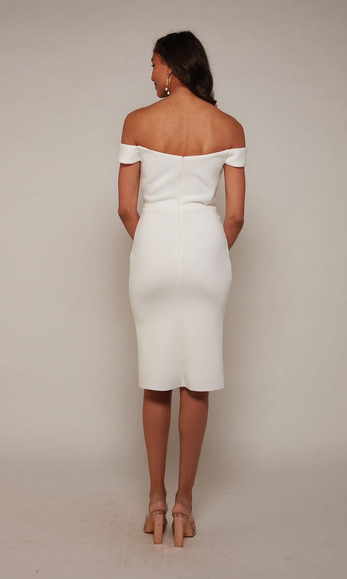 A woman faces away, wearing the Alyce 70047 knee length semi-formal dress in white with nude heels, set against a plain light background.